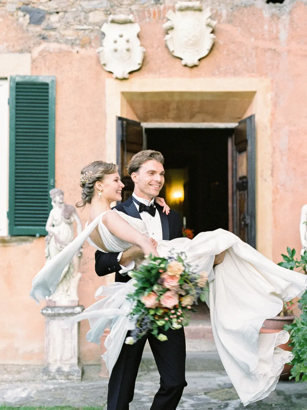 A fairytale moment unfolds as the newlyweds. Groom carrying bride holding her in his arms in the courtyard of a stunning medieval villa in Tuscany