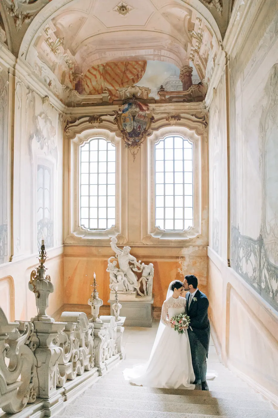 Beautiful wedding couple stand on the majestic staircase with frescoes on the wall and ceiling in the old Italian villa Arconat
